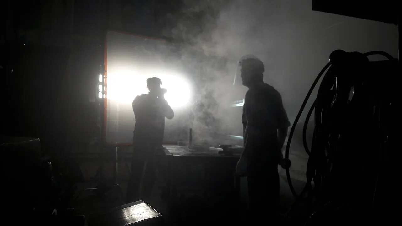 Two people silhouetted against a bright light in a hazy workshop.